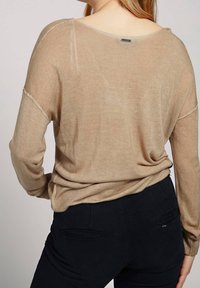 Suéter color marrón claro en suave tejido de punto con mangas largas, un escote amplio y puños de canalé, combinado con pantalones negros con detalles de bolsillos.