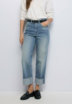 Mujer vestida con jeans azul claro con puños, cinturón negro, camiseta blanca de manga larga y mocasines negros, de pie frente a un fondo blanco sencillo.