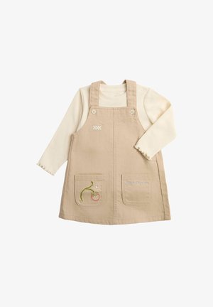 Beiges ärmelloses Kleid mit Fronttaschen und gestickten Details, getragen über einem langärmeligen cremefarbenen Hemd mit gewellten Manschetten.