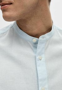 Camicia azzurro chiaro con colletto e scollo con bottoni. Realizzata in un tessuto testurizzato, presenta una leggera lucentezza e un design pulito e minimalista.