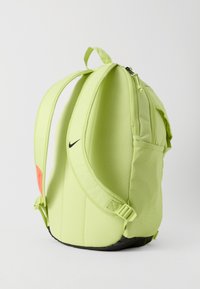 Mochila Nike verde claro con correas acolchadas para los hombros, bolsillo lateral y suela negra, mostrada desde un ángulo lateral sobre fondo blanco.