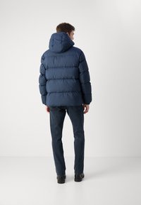 Marineblå puffjakke med hætte, der har et quiltet design og elastiske manchetter. Båret over mørke jeans og sorte sko, set fra ryggen.