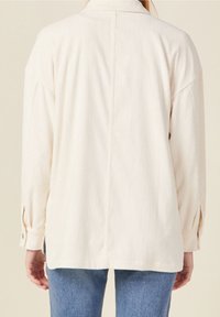 Cream corduroy shirt with a relaxed fit, featuring long sleeves, a collared neck, and button accents on the cuffs and side vents.