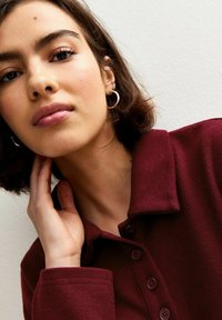 Camicia rosso borgogna con un tessuto strutturato e colletto. La modella si tocca il collo, indossando piccoli orecchini a cerchio in argento su uno sfondo semplice.