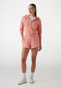 Roze zip-up hoodie met textuur logo en bijpassende shorts. Gemaakt van comfortabele stof, met zakken en een relaxte pasvorm. Witte sneakers.