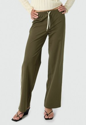 Žena oblečená v olivovo zelených širokých nohaviciach s bielym šnúrkou a krémovým svetrom, stojaca s rukami v bok, obutá v čiernych sandáloch na klinovom podpätku.