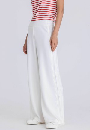 Personne portant un pantalon blanc à jambes larges, des baskets blanches et un haut à manches courtes rayé rouge et blanc, debout devant un fond clair uni.