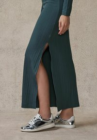 Abito maxi a coste color teal con spacco anteriore, abbinato a sneakers metalliche argento con lacci e dettagli bianchi, su uno sfondo testurizzato.