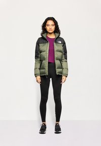 Polstret jakke med grønn kropp og svarte ermer, glidelås lukking, sidelommer, kombinert med en lilla topp, svarte leggings og svarte joggesko.