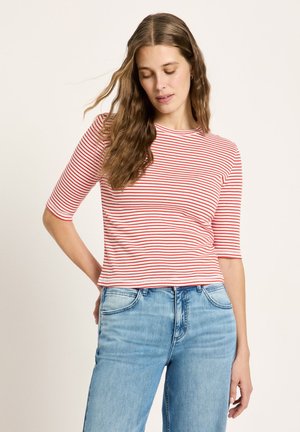 Woman with long wavy hair wearing a red and white striped half-sleeve shirt and light blue jeans, standing against a plain background.