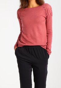 Top de manga larga rosa con detalles de encaje en los hombros, combinado con pantalones negros de cintura elástica. Textura suave y diseño ajustado.