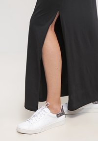 Zwarte maxi-jurk met een zijscheur, gedragen met witte sneakers met een gestructureerde grijze accent op de hiel. Glad stof en vloeiend ontwerp.