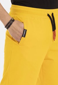 Geel joggingbroek met een elastische tailleband en zwarte trekkoord. Voorzien van zijzakken en een label "CIPOG&AXX" op de rand van de zak.