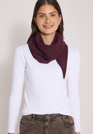 Jeune femme aux longs cheveux foncés portant une écharpe en laine bordeaux, une chemise blanche à manches longues et un pantalon marron taille haute, souriant à la caméra.