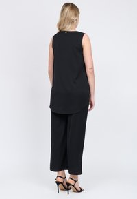 Top senza maniche nero con orlo curvo e un piccolo dettaglio dorato sul retro, abbinato a pantaloni neri a gamba larga e sandali neri con tacchi alti.