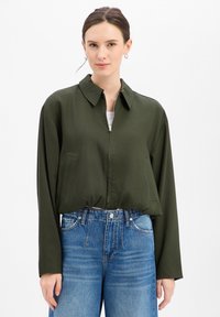 Olijfgroene cropped jas met een kraag, ritssluiting aan de voorkant en zijzakken. Gestyled met een high-waisted blauwe jeans met een relaxed fit.