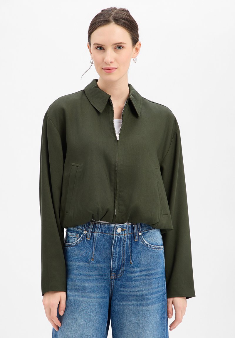 Olijfgroene cropped jas met een kraag, ritssluiting aan de voorkant en zijzakken. Gestyled met een high-waisted blauwe jeans met een relaxed fit.