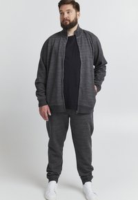 Mörkgrå träningsset med en zip-fodrad jacka med hög krage och matchande byxor. Båda delarna har en strukturerad yta. Vita sneakers.