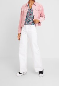 Giacca cropped in velluto a coste rosa, top floreale a scollo a V, jeans bianchi a gamba larga e sneaker nere. Evidenti contrasti di materiali, varietà di colori e texture.