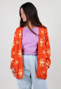 Cardigan orange vif avec des motifs abstraits crème, doté de boutons sur le devant. Associé à un t-shirt lavande et un jean bleu clair.