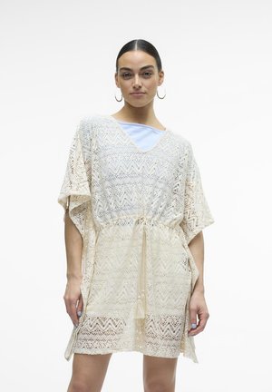 Abito coprente in pizzo beige con scollo a V, maniche corte, vita arricciata e dettaglio con frange, caratterizzato da un motivo a zigzag su tutta la superficie.