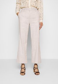 Žena ve béžových širokých kalhotách, vyšívané krémové halence a černých sandálech na podpatku, stojí se rukama v kapsách.