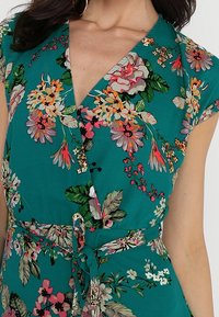 Robe portefeuille teal à manches courtes et ceinture nouée, ornée de fleurs roses, blanches et oranges, ainsi que de feuilles vertes.