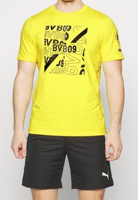 Camiseta de algodón amarilla con un diseño gráfico negro que incluye texto y formas. Combinada con pantalones cortos negros que exhiben un logotipo de Puma.