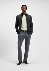 Mörkgrå zip-up tröja, krämfärgad polo-skjorta och ljusgrå byxor. Svart sneakers med vita sålar. Modellen står med händerna i fickorna.