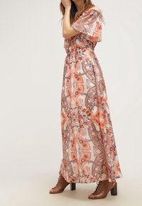 Kobieta w sukience do ziemi, bez ramion, w kwiatowy wzór w pomarańczowe i brązowe motywy, nosząca brązowe sandały na obcasie z wiązaniem.