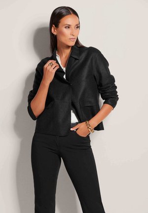 Schwarze, kurz geschnittene Jacke aus einem weichen Stoff, mit einem Kragen, zwei Fronttaschen und Dreiviertelärmeln. Trägt man über einem weißen Top und schwarzen Hosen.