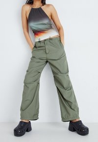 Czarny top na halter z gradientowym wzorem kolorystycznym, oliwkowe workowate spodnie, oraz czarne klompy z wycięciami. Modelka stoi w neutralnym otoczeniu.