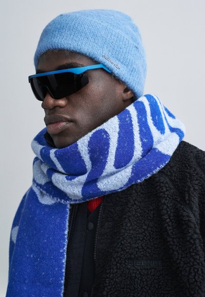 Bărbat purtând o căciulă tricotată bleu deschis, fular albastru cu alb cu model, geacă neagră pufoasă și ochelari de soare negri cu rame albastre, privind într-o parte.