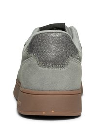 Grå mockaskor med en texturerad mörkgrå läderdetalj vid hälen och en brun gummisula. Har en sydd söm längs hälen.