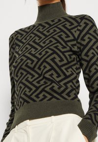 Olivgrüner Pullover mit einem schwarzen geometrischen Muster, hohem Kragen, gerippten Bündchen und Saum, gefertigt aus einem gestrickten Stoff.