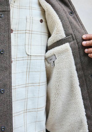 Braune Woll-Außenjacke mit beigem Sherpa-Futter. Das Innere verfügt über ein cremefarbenes kariertes Hemd mit hellbraunen Linien und einer Brusttasche.
