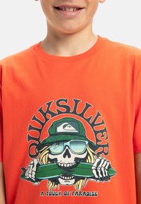 Oranžové bavlněné tričko s potiskem lebky v brýlích a zeleným objektem, s textem "Quiksilver" a kruhovým designem.