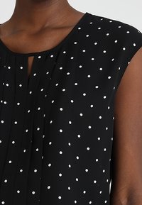 Blouse sans manches noire à pois blancs, avec un col rond et une petite fente à l'avant, ainsi que des plis verticaux.