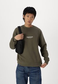 Sudadera verde oliva con el texto "Originals Studio", combinada con un pequeño bolso cruzado negro. Jeans azules con un detalle de costura visible.