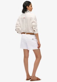 Shorts en denim blanc avec des ourlets effilochés, associés à une chemise en lin légère à boutons. Une ceinture tressée marron souligne la taille. Sandales plates marron.