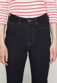 Jeans de mezclilla oscura con una cintura alta, botón de cobre y costuras naranjas. Llevados con una camisa de manga larga a rayas rojas y blancas.