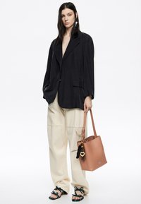 Blazer nero oversize, pantaloni crema a gamba larga, sandali neri decorati e una borsa a tracolla in pelle pesco con charm decorativi.