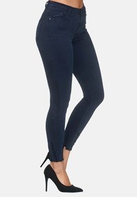 Jeans ajustados de color azul oscuro con detalle de dobladillo deshilachado, fabricados con tejido de mezclilla elástica. Diseñados con una cintura de altura media y estilo de cinco bolsillos.