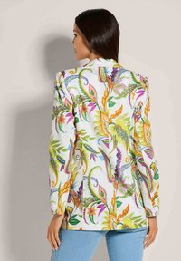 Frau mit langen dunklen Haaren, die ein weißes Blazer mit bunten Blumen- und Paisleymustern trägt, kombiniert mit hellblauen Jeans und mit dem Rücken zum Betrachter.