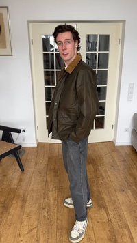 Νέος άνδρας που φορά καφέ μπουφάν με γιακά ταμπά, γκρι τζιν και λευκά αθλητικά παπούτσια, στέκεται μέσα σε εσωτερικό χώρο σε ξύλινο πάτωμα κοντά σε πόρτα με γυάλινο πάνελ.