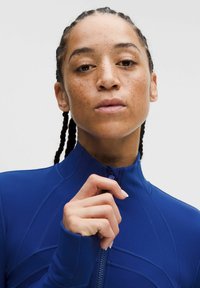 Haut de sport bleu avec col montant et fermeture éclair, présentant des motifs texturés et une coupe ajustée, fabriqué dans un matériau lisse et extensible.