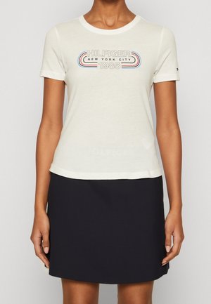Femme portant un t-shirt blanc à manches courtes avec le texte "HILFIGER NEW YORK CITY 1985" et une jupe noire.