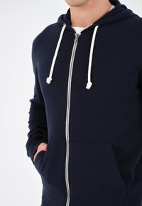 Mornarska zip-up hoodica s bijelim vezicama, prednjim džepovima i mekim materijalom. Sadrži metalni zip i uske rukave.
