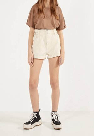 Person iført en brun kortærmet skjorte, højtaljede cremefarvede shorts med knapper og sorte høje sneakers med hvide snørebånd.