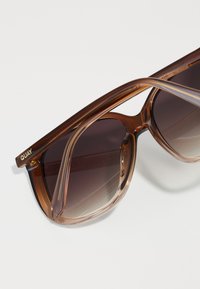 Braune, durchsichtige Sonnenbrille mit grau verlaufsfähigen Gläsern, breiter rechteckiger Form und goldener "QUAY"-Markierung am Bügel.